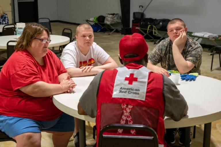 woman and her sons sit at a table in a Red Cross shelter with a volunteer
