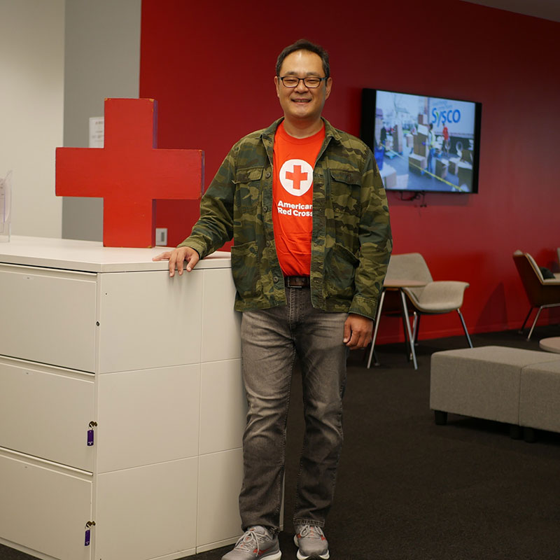 Sung Hwang standing next to Red Cross logo on top of a file cabinet, wearing a Red Cross shirt.