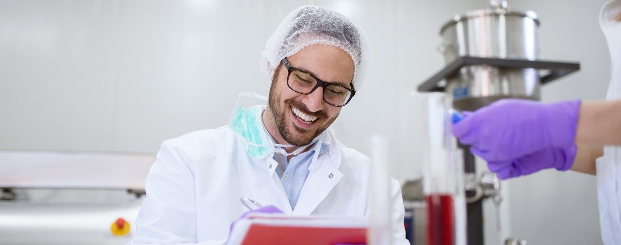 Man wearing, glasses hair net, rubber gloves, and white coat writing on cli[pboard and smiling.