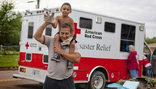 Man with child on his shoulders next to a Red Cross Disaster Relief vehicle