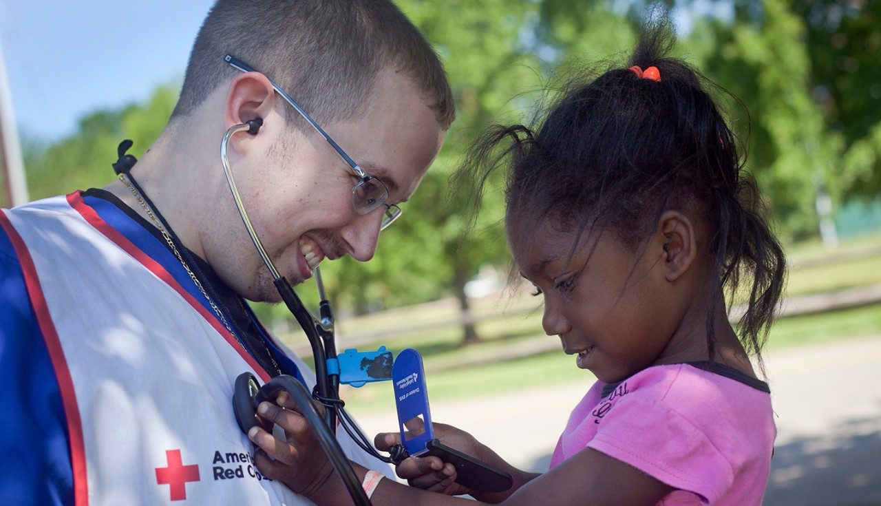 Red Cross nurse with stethoscope holding child who is using stethoscope on nurse