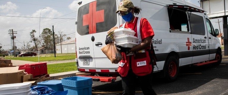 Red Cross volunteer holding packaged food next to a Red Cross vehicle.
