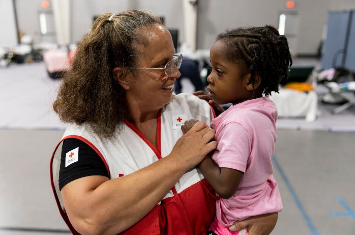 Female volunteer in Red Cross vest holiding a toddler in a shelter