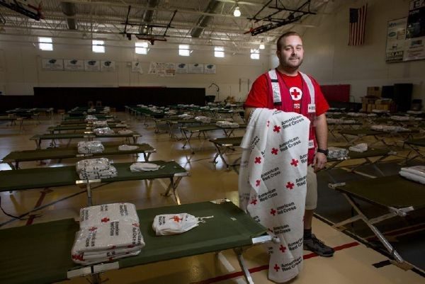 Red Cross volunteer holding a Red Cross blanket and standing in a shelter surrounded by shelter cots 