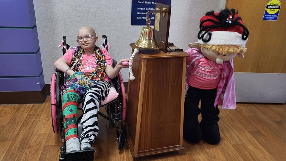 Girl in wheelchair ringing bell with large doll standing next to bell