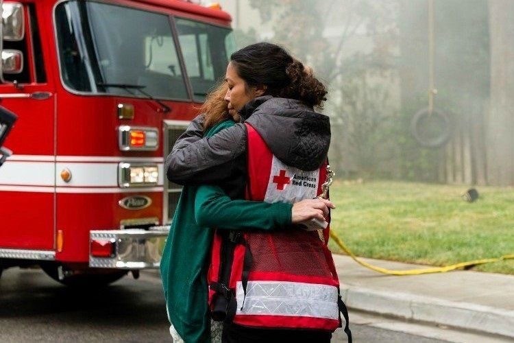 Red Cross volunteer in red vest hugging a woman in front of a fire truck