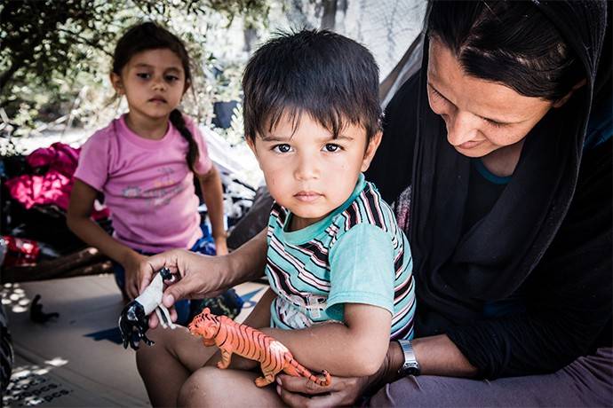 Greece: Lesbos, 21 August 2015   Suhaida and her sun Mahdi. In the background is her daughter Mahdia. Both these names mean 'rightly guided'. The toys the children play with were bought for them by their parents in Iran, and they have carried them with them all the way to Greece. The family is from Afghanistan, and are waiting to be able to register on Lesbos before seeking a safer, happier life.   Each day, thousands arrive on Greek shores seeking safety, security and a future. Hundreds of people have been arriving on the island of Lesbos. The Red Cross is providing assistance to those passing through Lesbos before they carry on with their journey.   Photo: Stephen Ryan / IFRC