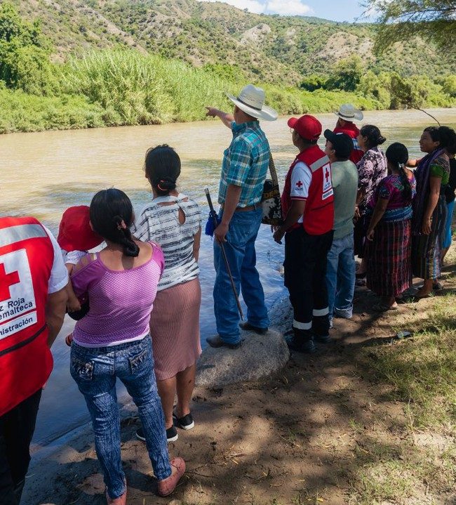 A group of locals and several Red Cross volunteers are looking out at a flooded river