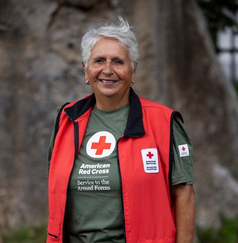 An elderly woman standing in front of a tree in Puerto Rico takes a portrait, in a green Red Cross Service to the Armed Forces t-shirt with a red Hero Care vest on top.