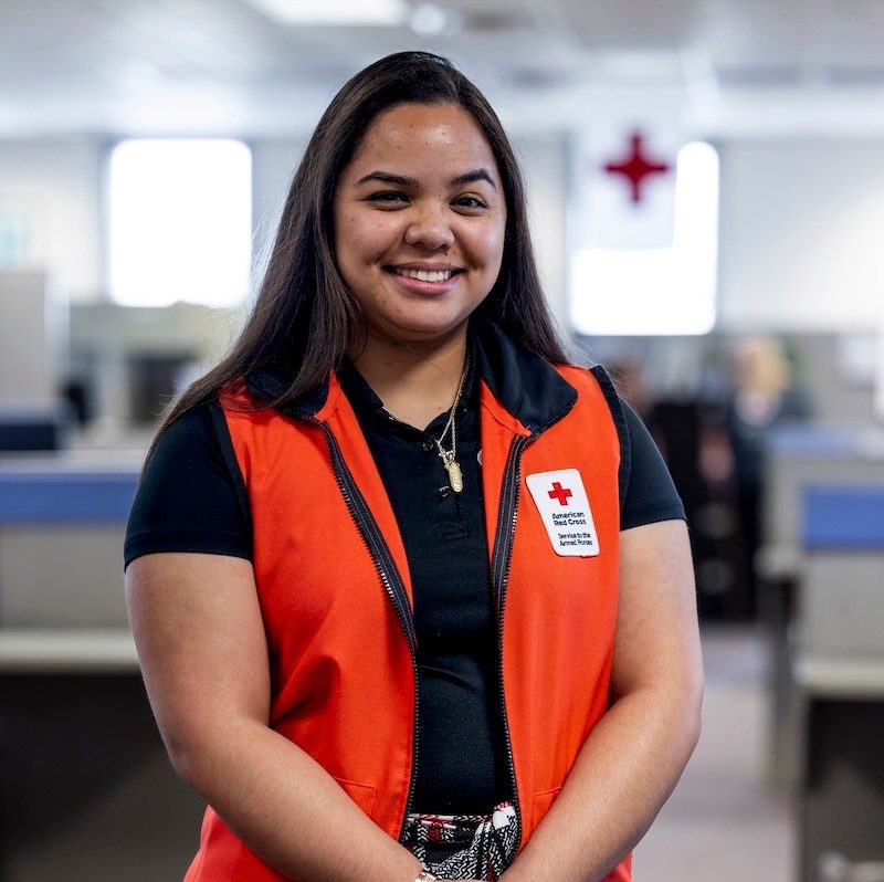 A young woman with shoulder-length dark hair stands in a Red Cross office wearing a Hero Care vest.