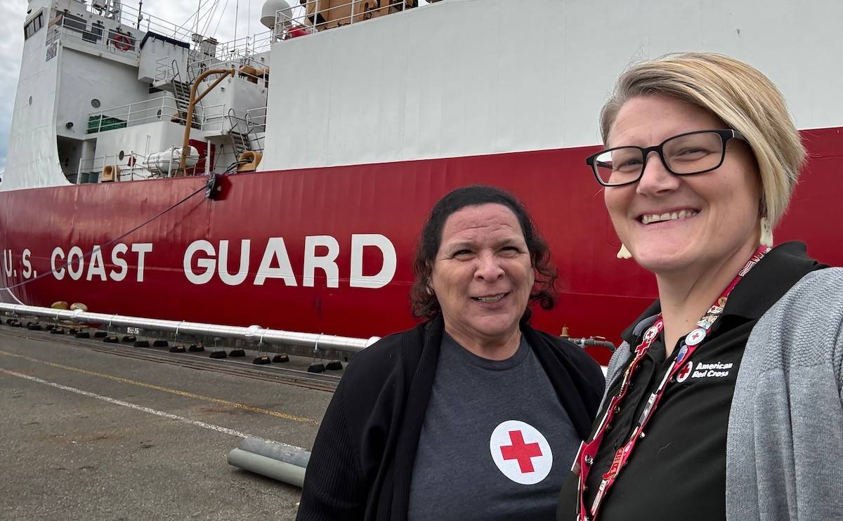 Two women in Red Cross gear stand in front of a US Coast Guard ship. 