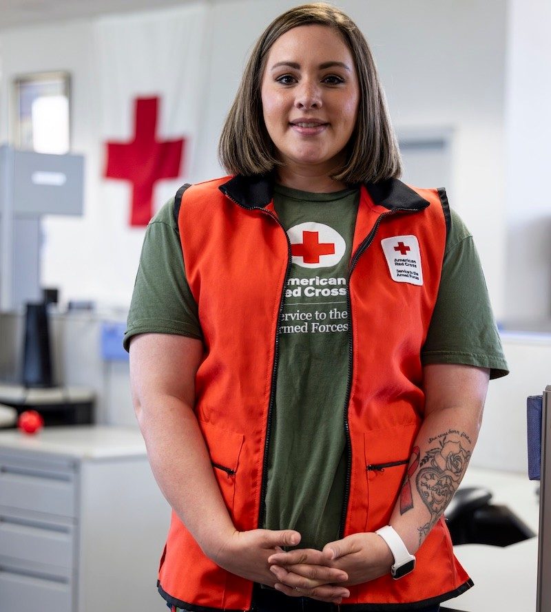 A young woman with a tattoo on her forearm and wearing a Red Cross Hero Care vest smiles for a portrait