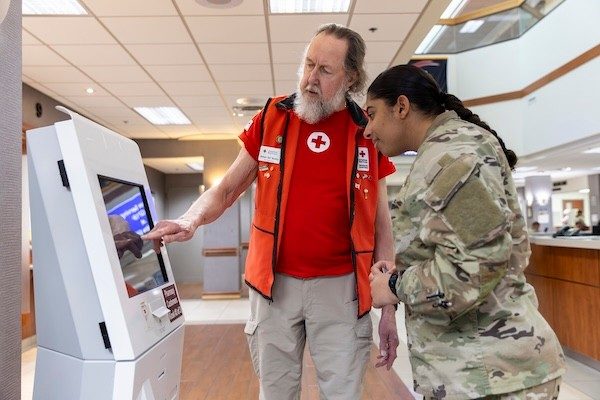 Red Cross volunteer wearing a Hero Care vest shows uniformed servicemember how to use a touchscreen in a lobby.