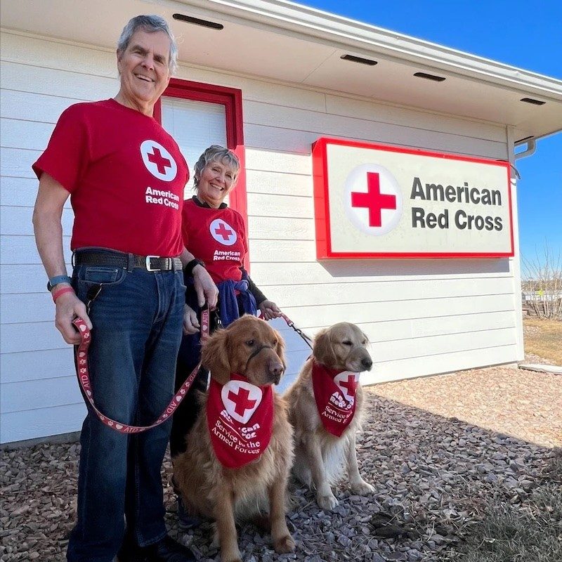 Two Red Cross volunteers are posing in front of a Red Cross office with their certified service dogs.