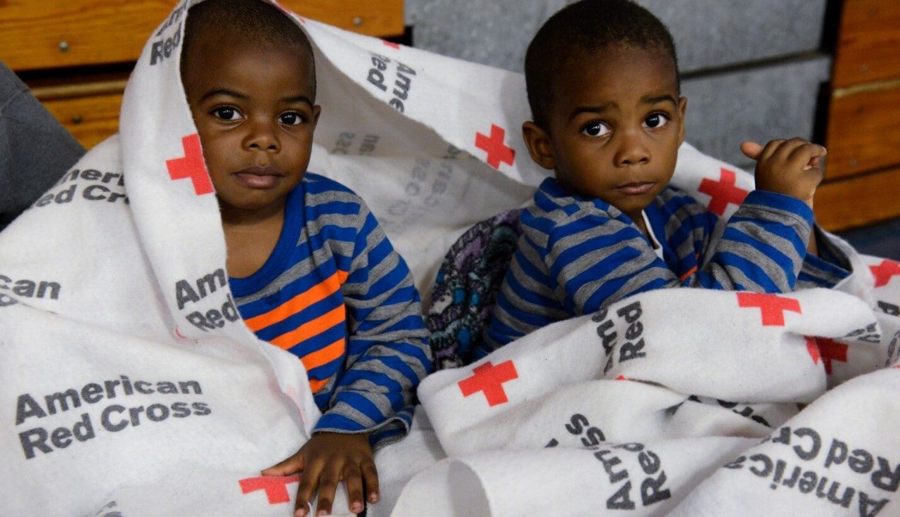 Two boys wrapped in a Red Cross blanket at a shelter.