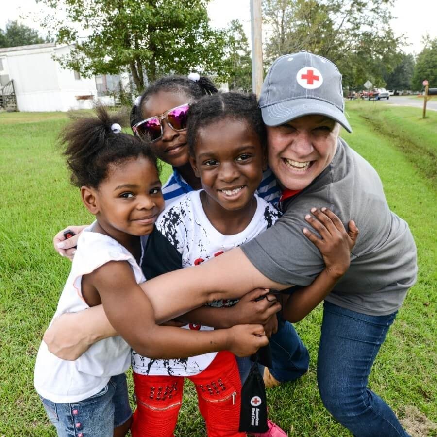 A smiling Red Cross volunteer hugging kids outside.