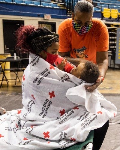 A woman holding her baby wrapped in a Red Cross blanket at a shelter.