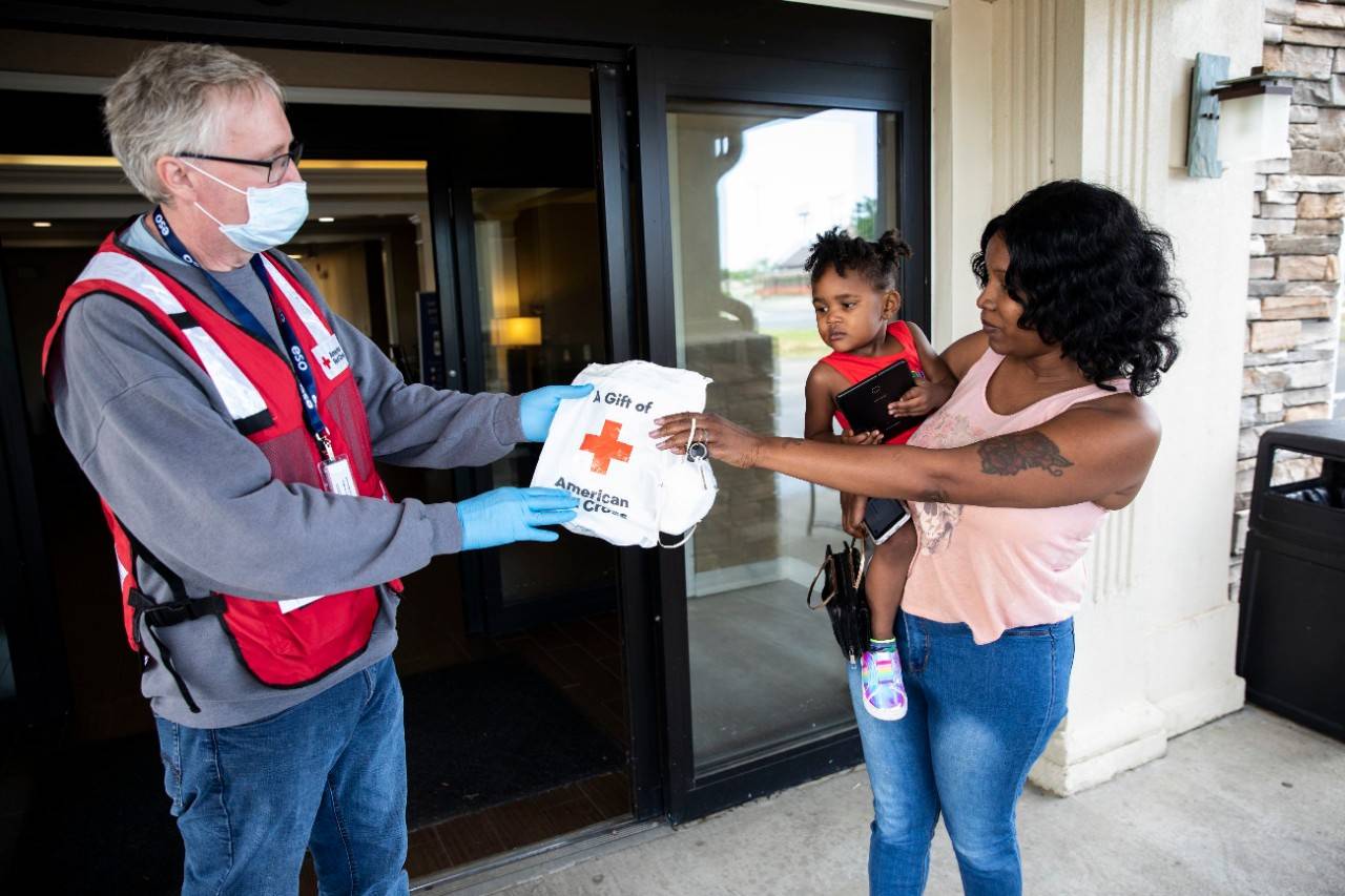 April 18, 2020.  Monroe, Louisiana.Charles "Harry" Murphy, Jr., BS, NRP the Emergency Management Coordinator for the Louisiana Region, Cap/West Chapter, hands a bag containing masks to Natechia and her daughter Nyla, 2 years old, whose home was damaged by a recent tornado in Monroe, LA, on Saturday, April 18, 2020. Neatechia and her family are one family of hundreds of people in Monroe who needed shelter after the storm. Normally the Red Cross would set up shelters, but due to the pandemic and social distancing guidelines, people who were unable to stay in their homes have been given rooms in various local hotels.Photo by Scott Dalton/American Red Cross
