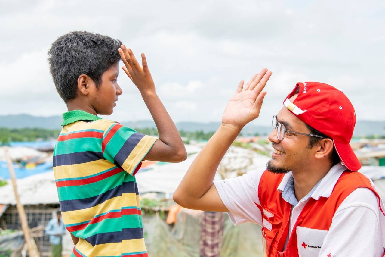 June 27, 2019. Cox’s Bazar, Bangladesh. Sofi Alam, age 8, high-fives American Red Cross staff member, Shariar Morshed Saad. Sofi is a force. He walks confidently through the winding hillsides of Kutupalong—a camp for displaced families in Cox’s Bazar—high-fiving friends and dodging auto-rickshaws on the muddy, slippery streets. Migrants like Sofi don’t have access to formal education, but he has chosen to attend English class at a nearby learning center every day. “I want to be a teacher when I grow up because I want to give education to kids and communities who don’t have it,” he attests. In a place where hope can feel hard to come by, Sofi personifies it. His favorite activities? “I love to play soccer and I love to learn.” Sofi doesn’t know the name, “Red Cross Red Crescent.” Instead, he recognizes the symbol. He says that kids in the camp call the red crescent and red cross emblem as they see it, “Moon and Star.” He knows that people wearing that emblem help to strengthen shelters in the camp and teach families how to tie down their roofs when storms hit. He also knows what kids should do if storms hit. “Kids shouldn’t play in the floods. They should go around the floods.” And “Kids should stay home during cyclones and storms.”Since August 2017, more than 700,000 people have fled Rakhine State, Myanmar to seek safety in Cox’s Bazar, Bangladesh. Many arrived injured, malnourished, and devastated. They speak of dangerous journeys—walking days on end to reach the border and losing touch with family members along the way. Once in Bangladesh, they crowd into camps on muddy hillsides and live in structures made of bamboo, plastic, cardboard and sometimes corrugated metal sheeting. Monsoon rains and dangerous cyclone seasons put migrants at risk of landslides, floods, and destructive wind. Despite harsh conditions—and because there’s no possibility of evacuating the displacement camp during cyclones—migrants are volunteering to help their neighbors during natural disasters and other emergencies. The American Red Cross has been working alongside global Red Cross and Red Crescent teams to provide lifesaving aid to the families. We are training migrants on first aid, early warning systems, and other skills so they can respond to rain, wind, flooding, landslides, and cyclones. As part of the Bangladesh government’s Cyclone Preparedness Program (CPP), the American Red Cross, Bangladesh Red Crescent, and IFRC are training thousands of camp residents and local (host) community members. In addition, the Red Cross supports disaster simulation drills and equips volunteers to warn fellow residents of danger via megaphones, a flag system, and even loudspeakers at mosques. Photo by Brad Zerivitz/American Red Cross. 