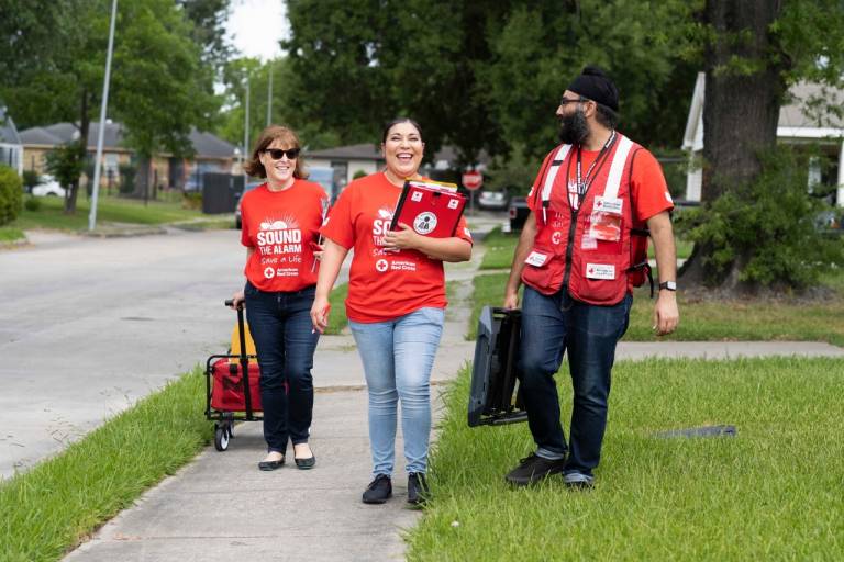 Three volunteers are walking down the street toward the camera wearing Red Cross tee shirts and carrying equipment. They are smiling