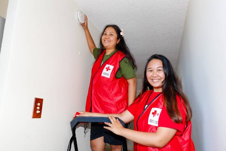Dos voluntarios con chalecos de la Cruz Roja trabajan en una escalera para instalar detectores de humo
