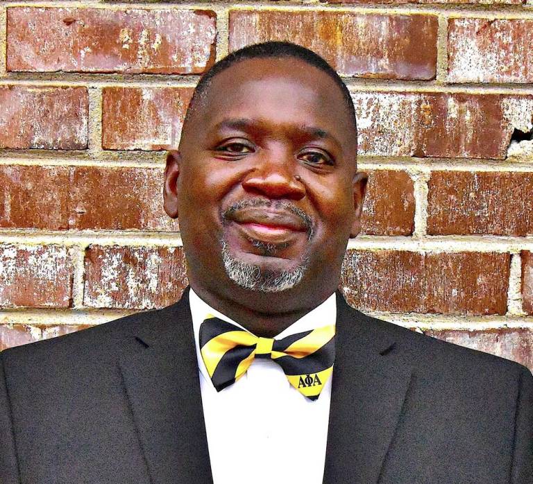 photo of Rubin, a black man standing in front of a brick wall wearing a yellow and black bow tie and tuxedo