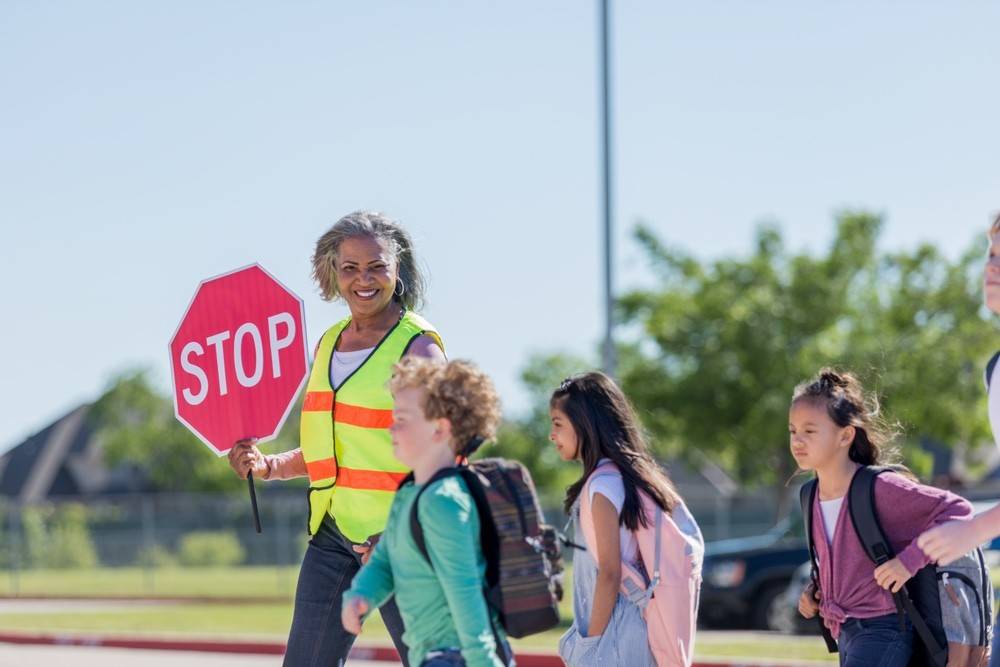 Helpful female crossing guard helps student cross a street on a crosswalk. The crossing guard is holding a stop sign.