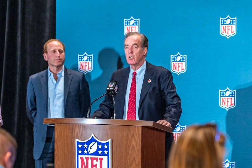 Jack McMaster of the Red Cross stands at a podium during a press conference. The podium and the wall behind have NFL logos on them.