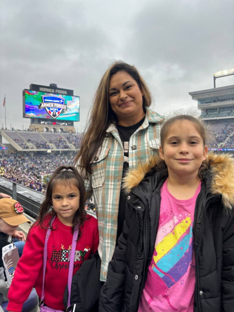 woman stands with her two daughters in the stands at a football game