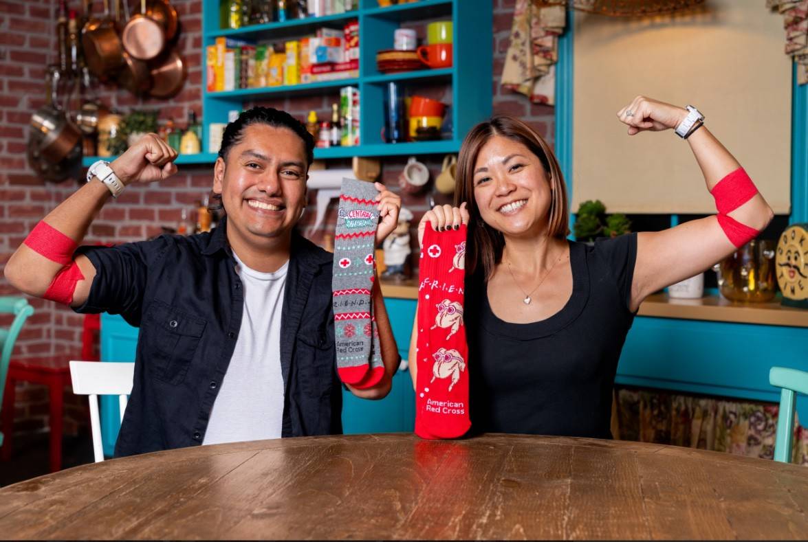 Dos recientes donantes de sangre están sentados en la mesa de la cocina, sonriendo y sosteniendo los calcetines de FRIENDS + Cruz Roja.