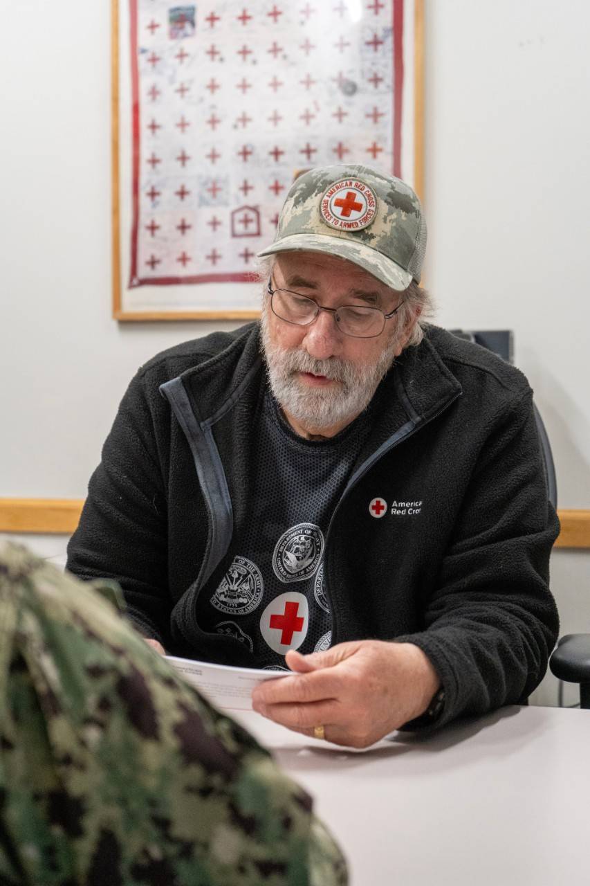 Un hombre con gafas, barba blanca y gorra de la Cruz Roja está sentado frente a un miembro del servicio militar mientras le lee un folleto.