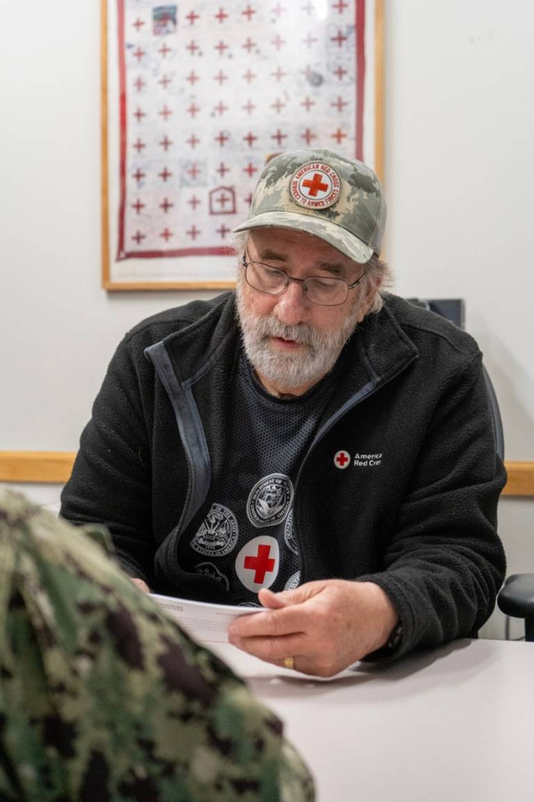 A man with glasses, a white beard and Red Cross hat sits across the table from a service member, reading them a flyer.