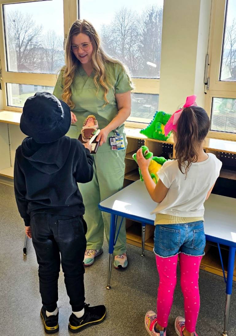A dental assistant in scrubs stands in front of two school-age children and demonstrates tooth cleaning techniques using a fake mouth.