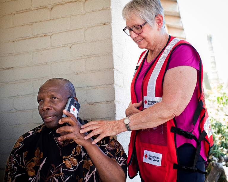 Woman in Red Cross vest looks on as man talks on a telephone.