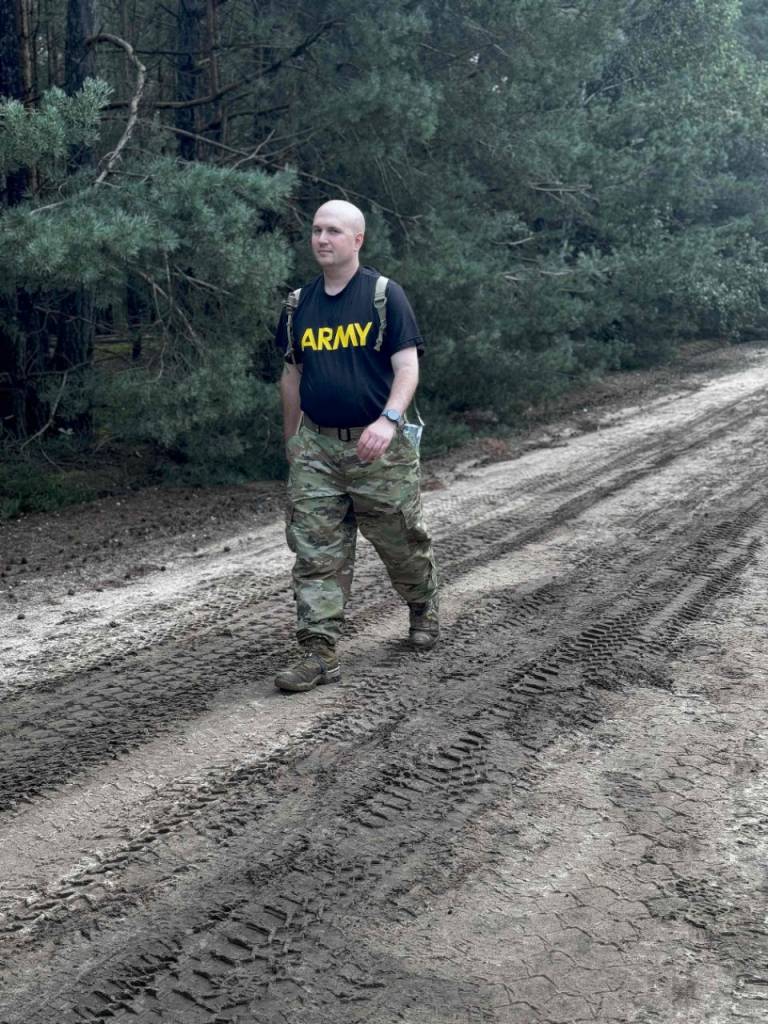 A soldier in a black t-shirt with “Army” in yellow letters across the chest, camo pants and boots marches through the mud carrying a backpack. 