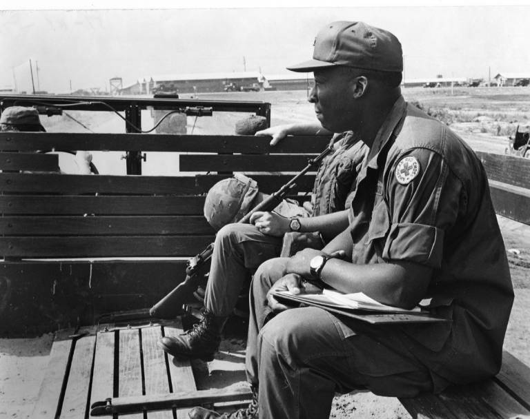 Black and white photo of two men sitting in back of truck in fatigues.
