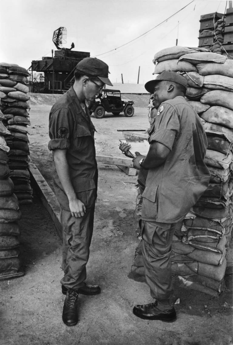 Black and white photo of two men behind piles of sandbags on military post