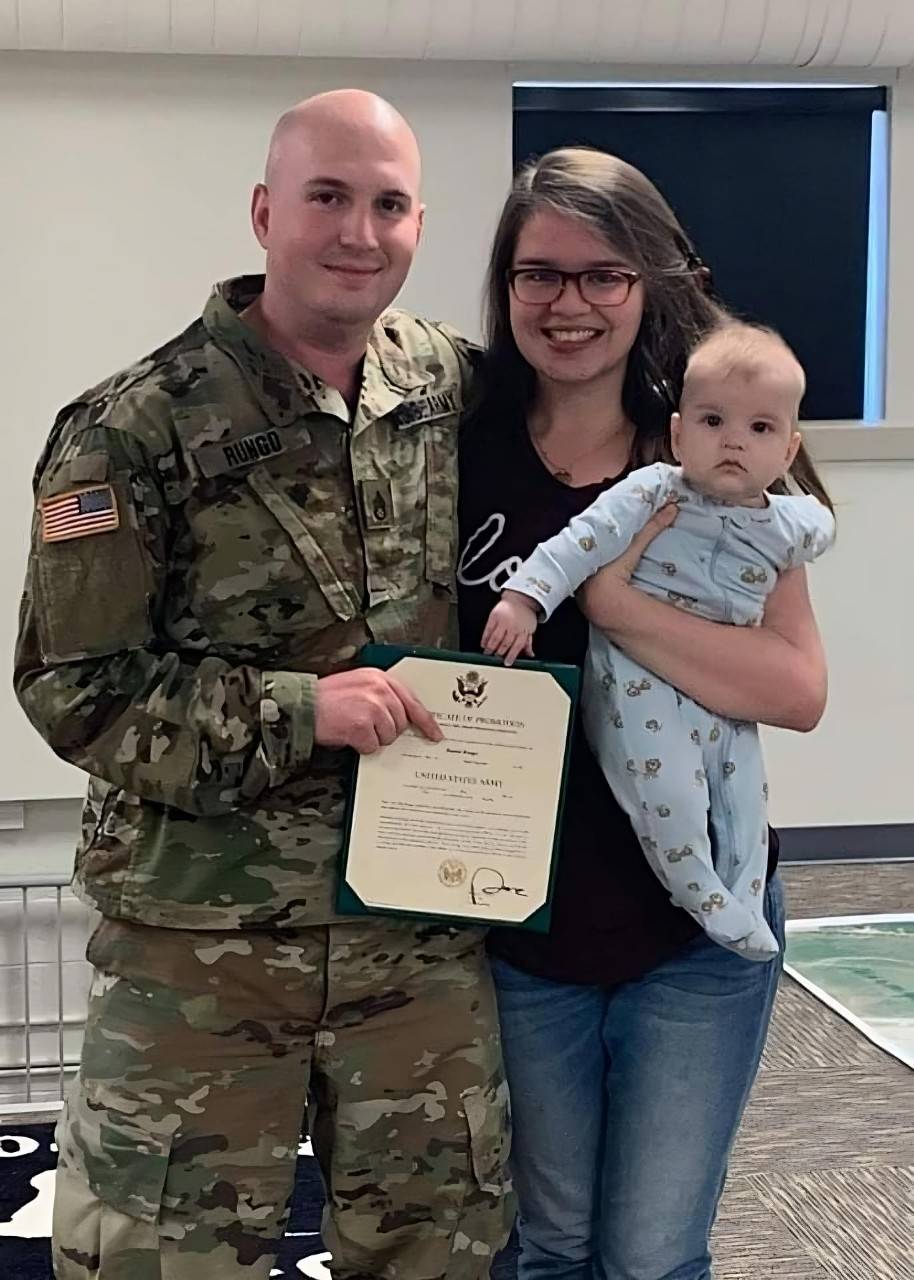 A man in an Army uniform holds his enlistment certificate and smiles with his arm around his wife. She cradles their baby.