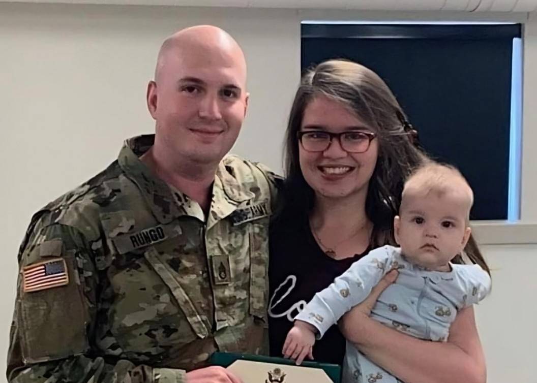 A man in an Army uniform holds his enlistment certificate and smiles with his arm around his wife. She cradles their baby.