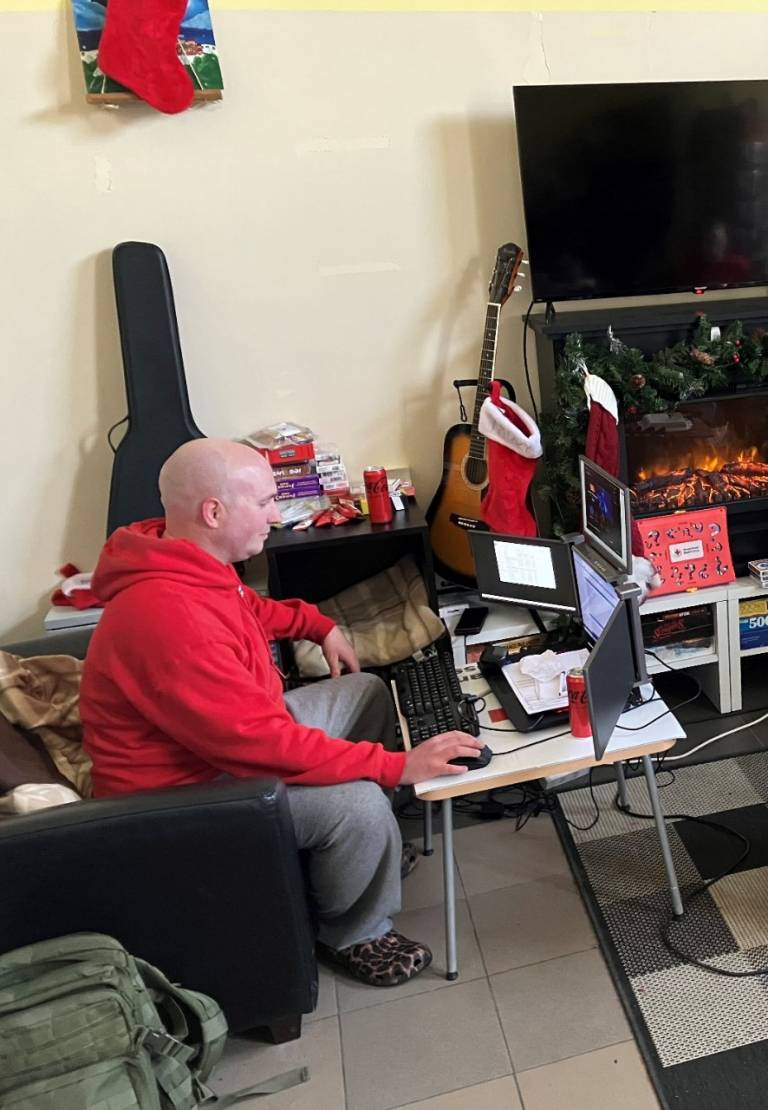 A bald-headed man with a Red Cross sweatshirt sits on a couch with a desk pulled up and works on the computer. The space is casual with a guitar, some games and festive decorations seen in the background.