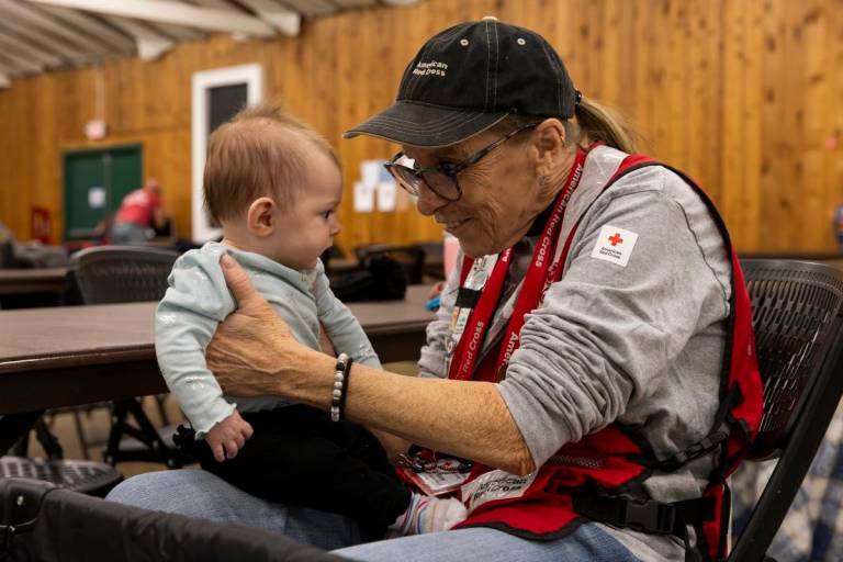 man in Red Cross vest interacts with infant in a shelter