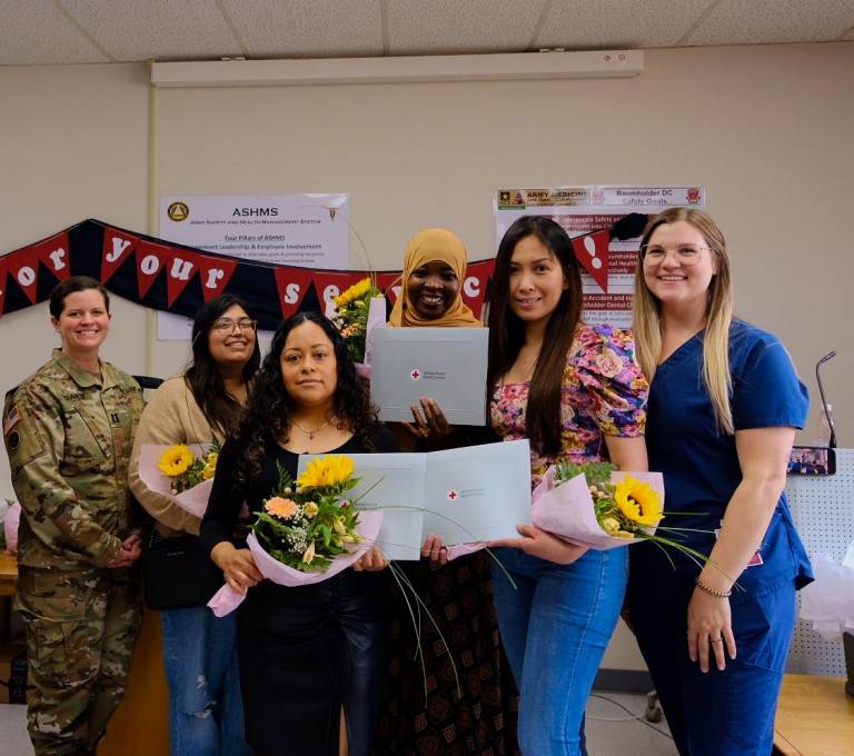 Four women proudly hold flowers and graduation certificates with a woman in an Army uniform and dentist in blue scrubs to their sides.