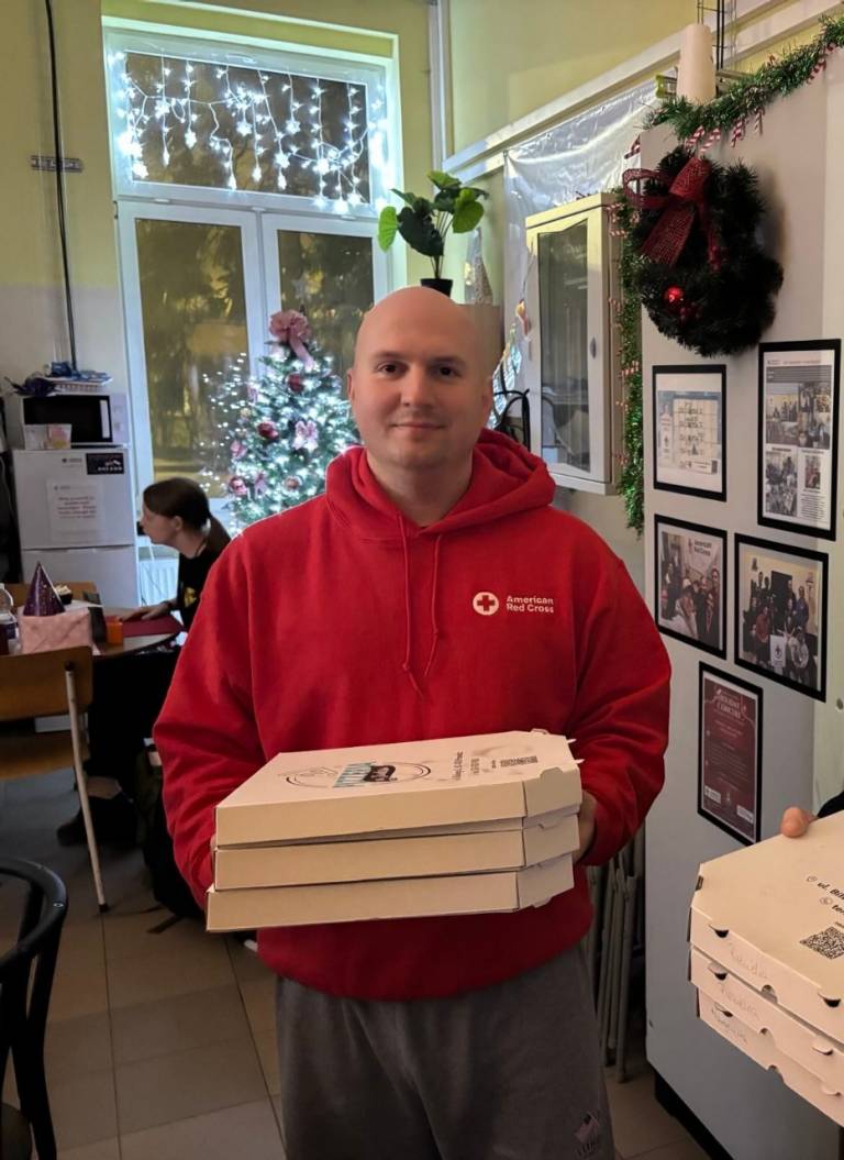 Red Cross volunteer wearing a Red Cross sweatshirt holds three pizzas standing beside a stack of others. Behind him are festive lights.