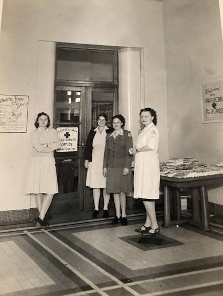 Cuatro mujeres en una foto en blanco y negro visten uniformes de enfermería antiguos y se encuentran dentro de un salón de mármol en la sede nacional de la Cruz Roja Americana.