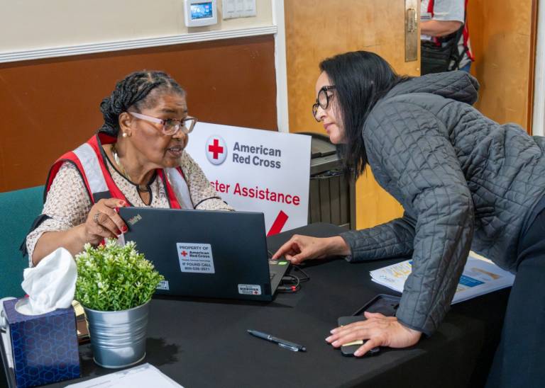 Woman in Red Cross vest shows laptop screen to woman standing next to the desk.