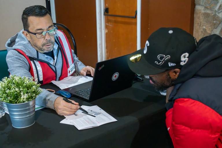 man in Red Cross vest sits on one side of a desk with another man in hooded sweatshirt sitting on the other side, both going over paperwork.