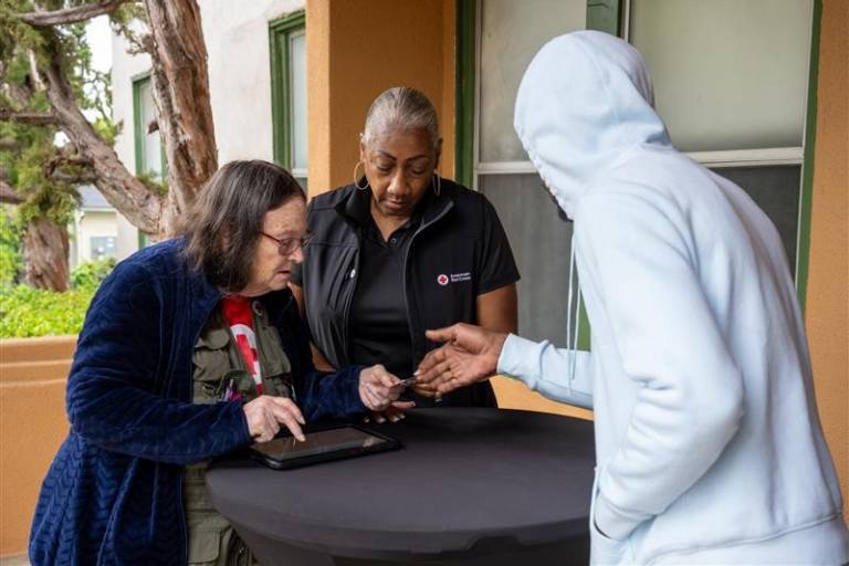 Two women identified as Red Cross workers stand on one side of a desk and man in a hooded sweatshirt stands on the other side as they go over paperwork.