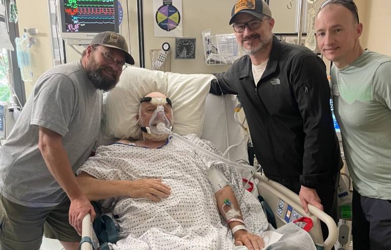 Kyle Zeck with his two brothers gather around a hospital bed with their father who wears a breathing apparatus following a triple bypass surgery.