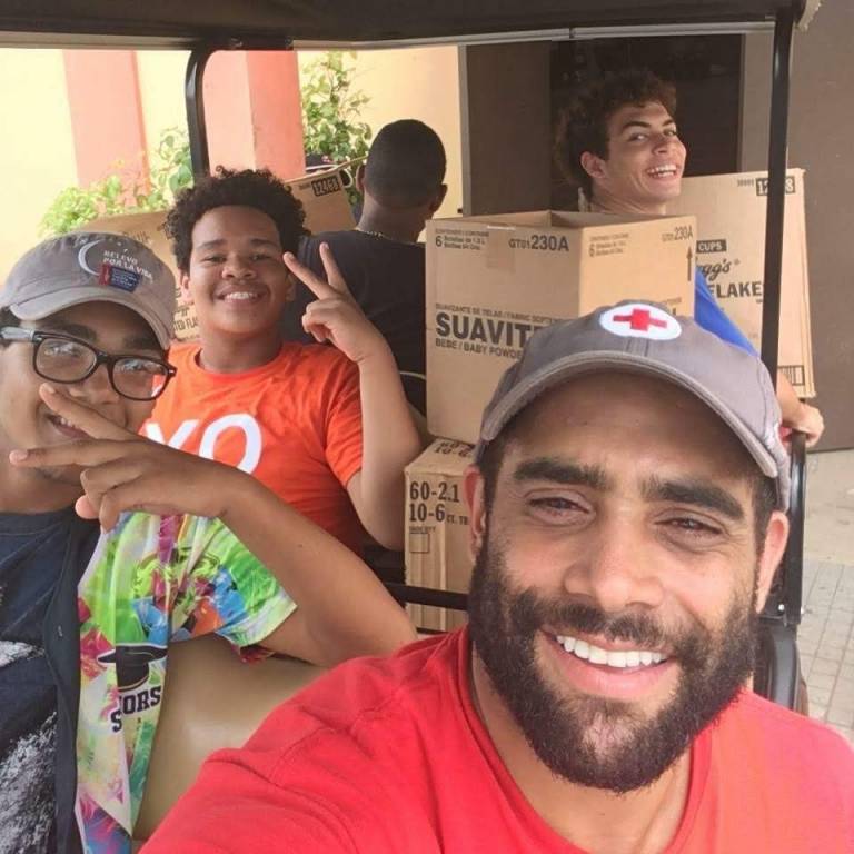 Shaun wears a Red Cross hat and takes a selfie with three youth volunteers, sitting in a golf cart filled with supplies. 