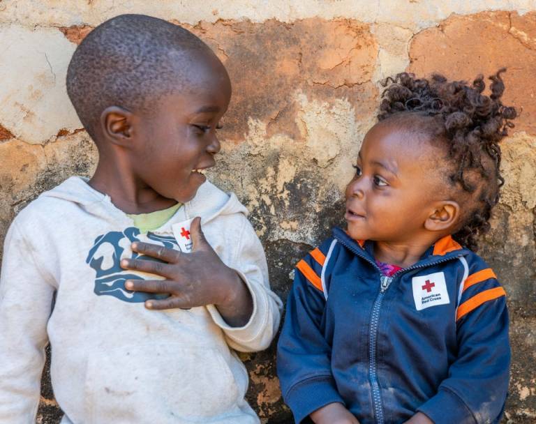 Dos niños pequeños con adhesivos de la Cruz Roja en sus chaquetas están sentados afuera